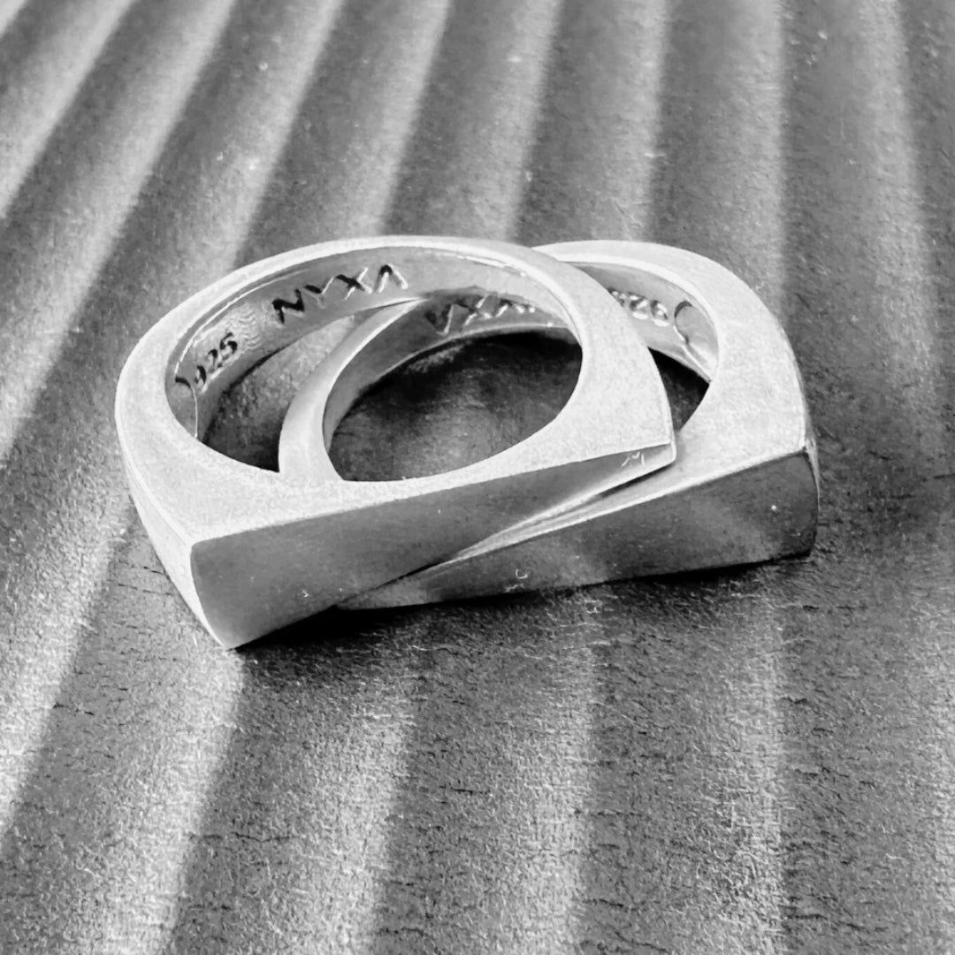 Close-up of two bold sterling silver rings with geometric design from the NYXA Vacation Jewelry Collection, resting on a textured black surface.